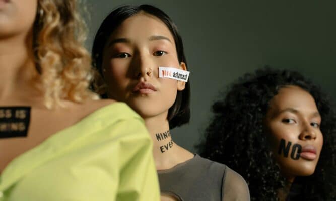 Three women stand in a line. The woman in center frame has a piece of paper on her face that reads "Not ashamed." A phrase in black on her chest reads "Bodily Integrity."