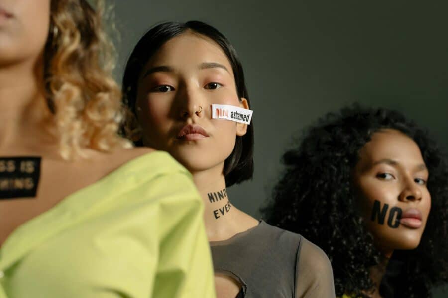 Three women stand in a line. The woman in center frame has a piece of paper on her face that reads "Not ashamed." A phrase in black on her chest reads "Bodily Integrity."