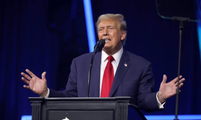 A man in a navy suit and red tie stands behind the podium with his arms outstretched.