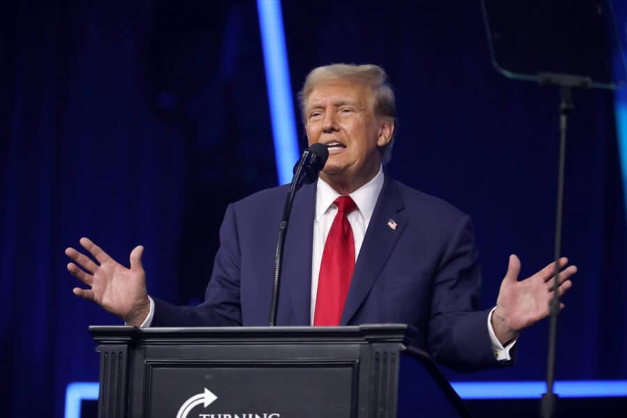 A man in a navy suit and red tie stands behind the podium with his arms outstretched.