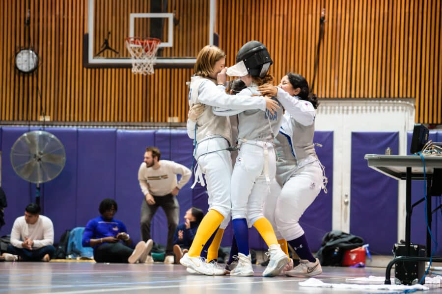 Fencers hugging and celebrating together on the gym floor after a match.