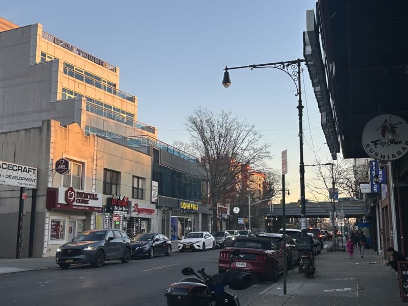 A large white building is in the left side of the frame, cast in golden light. A sidewalk is on the right side. Parked cars line both sides of the street. 