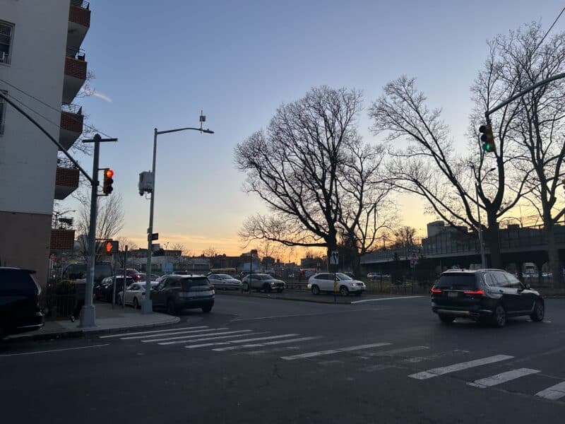 The photo is of a dark street lined with trees in the background. Orange light peeks out through the trees. 