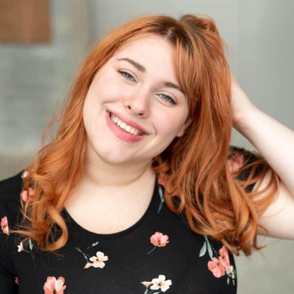 A woman with red hair in a black, floral t-shirt is in the center frame. She is smiling and has her hand in her hair.