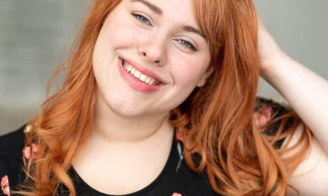 A woman with red hair in a black, floral t-shirt is in the center frame. She is smiling and has her hand in her hair.