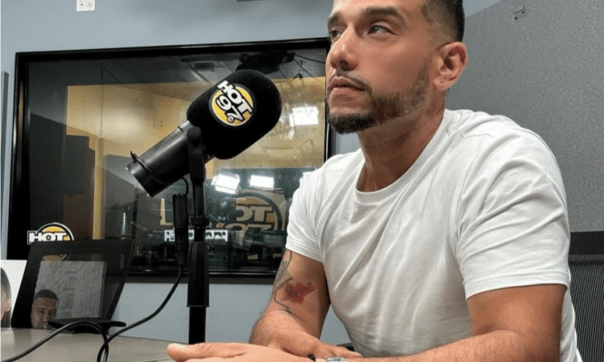 A man with a silver watch and white T-shirt looks off into the left of the frame. He sits at a table with a microphone in front of him.