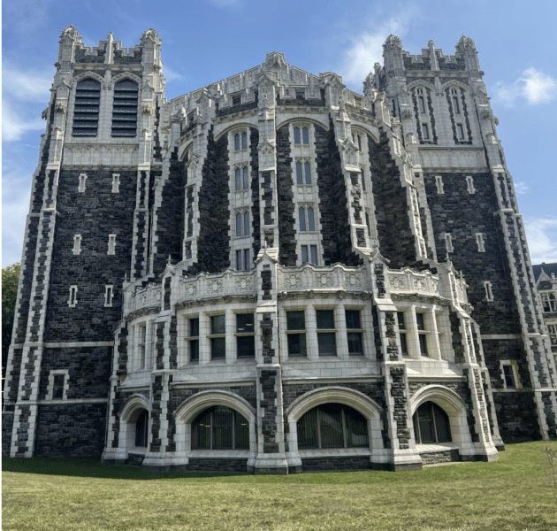 A gothic-style building with grey bricks and white trim stands on a green lawn. The blue sky peeks over the top of the building. 