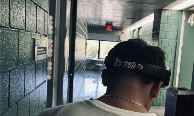 A student with headphones stands just to the right of the center of the frame in a green hallway. Only the back of their head is visible. Their headphones have a sticker that reads "$ad Boys."