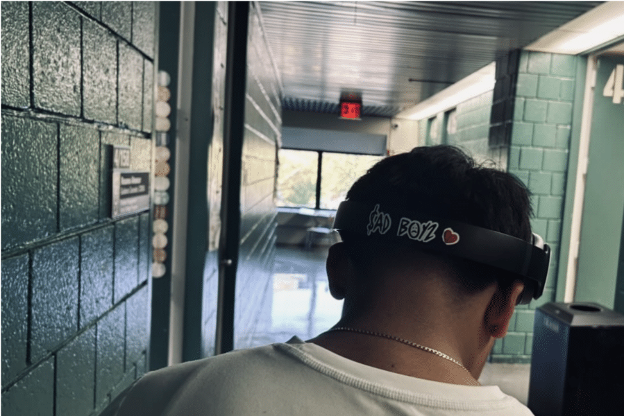 A student with headphones stands just to the right of the center of the frame in a green hallway. Only the back of their head is visible. Their headphones have a sticker that reads "$ad Boys."