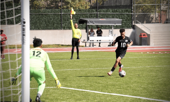 Three figures are on a green soccer field. Someone in a black uniform is in the right frame with the ball. Another figure is wearing green and is guarding the goal. A referee holds a yellow flag in the middle of the two players.