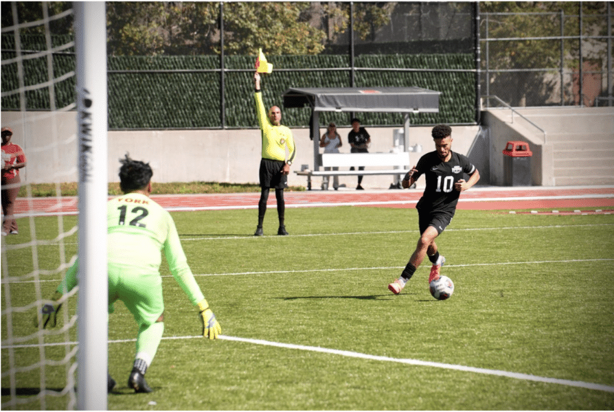 Three figures are on a green soccer field. Someone in a black uniform is in the right frame with the ball. Another figure is wearing green and is guarding the goal. A referee holds a yellow flag in the middle of the two players.