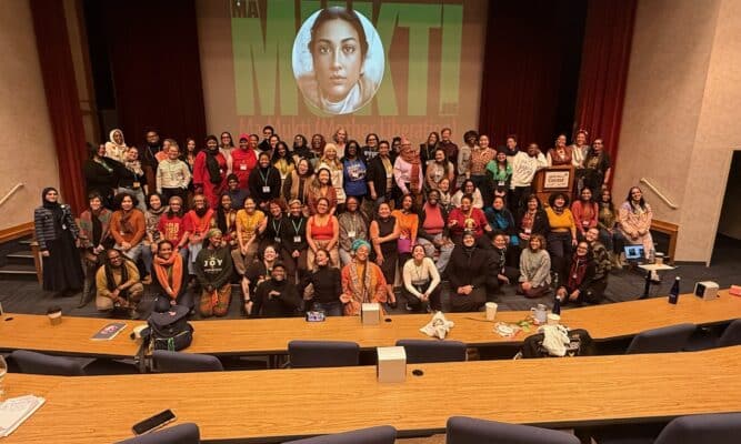 Women crowd together on a stage for a photo. There is a projection screen in the background with a woman's face.