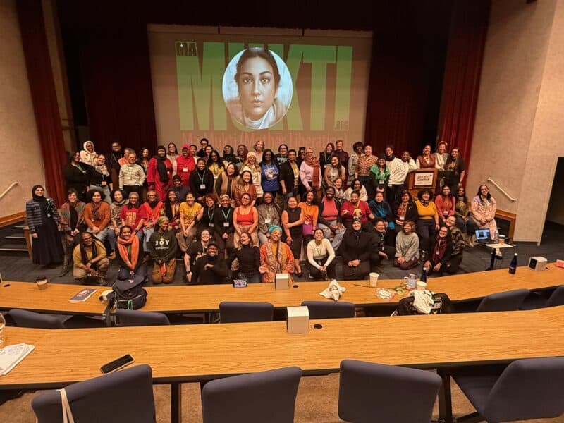 Women crowd together on a stage for a photo. There is a projection screen in the background with a woman's face.