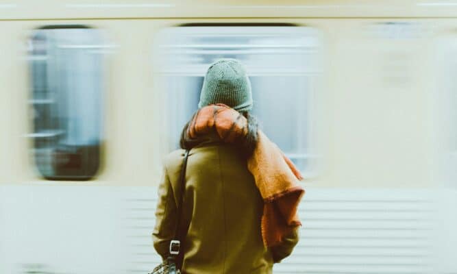 A woman in a hat and coat stands in beside a blurry, moving train car.