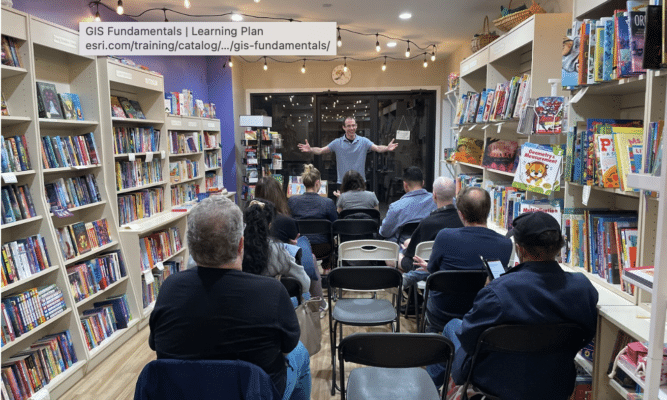 A man stands in the middle of the frame with his arms outstretched. People sit in folding chairs in front of him, with their backs toward the camera.