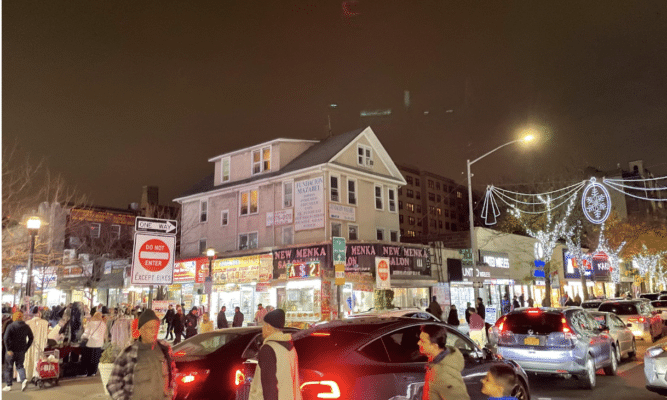 People cross at the crosswalk in Jackson Heights. Four people are walking in the foreground of the photo, cast in the red light of a car's tail light. A large grey house and a busy street corner are in the background.