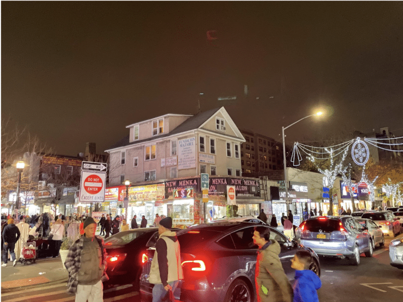 People cross at the crosswalk in Jackson Heights. Four people are walking in the foreground of the photo, cast in the red light of a car's tail light. A large grey house and a busy street corner are in the background.