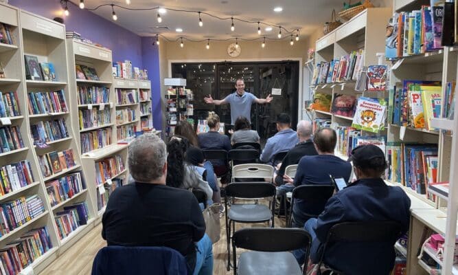 A man stands at the front of a room in a bookshop. There's three rows of people sitting facing the man, with their backs to the camera. They are surrounded by bookshelves and fairylights.