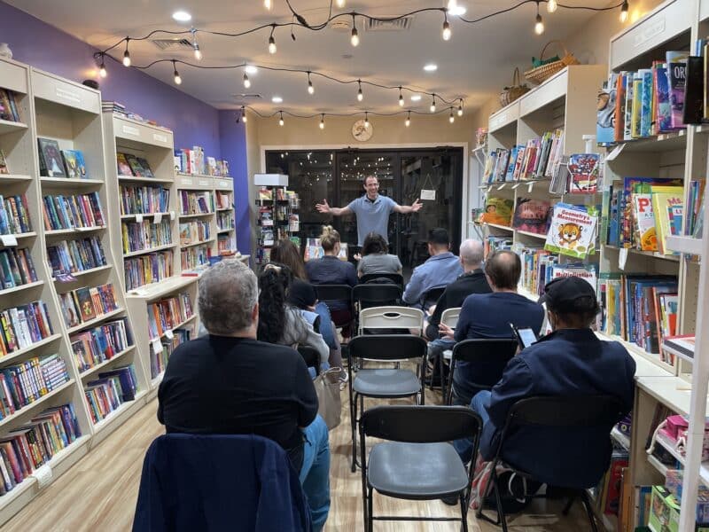 A man stands at the front of a room in a bookshop. There's three rows of people sitting facing the man, with their backs to the camera. They are surrounded by bookshelves and fairylights.