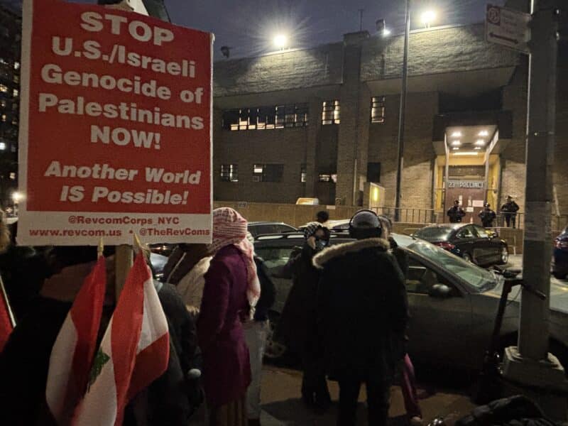 A red sign reading "Stop U.S/Israeli Genocide of Palestinians Now! Another world is possible!" is on the left side of the frame. Protestors stand on the sidewalk opposite from the 23rd precinct.