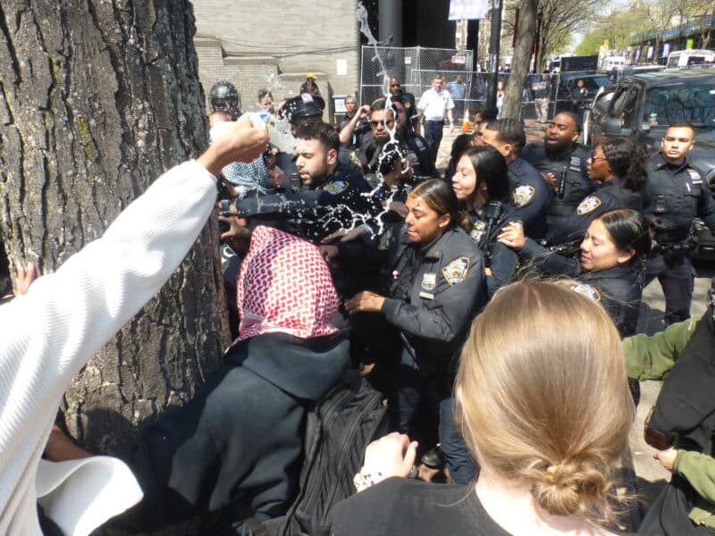 A crowd of students are in the foreground of the photo, with their backs toward the camera. Police are in the background, with their faces toward the camera. Someone with a plastic water bottle sprays water.