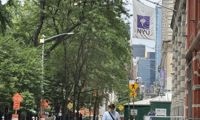 a man in a suit walks under a New York University flag on the sidewalk.