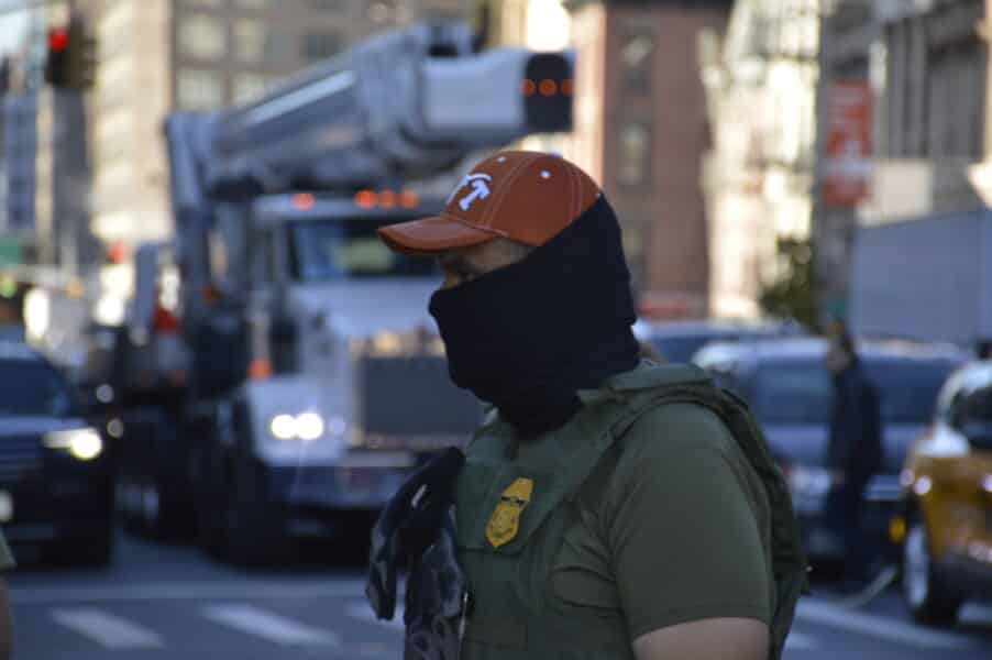 A masked ICE agent stands guard in front of ongoing traffic
