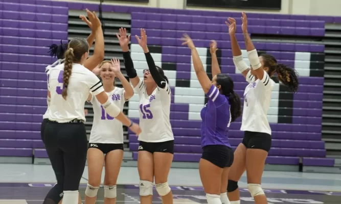 Group of college-aged volleyball players reach for a high face in celebration after a victory.