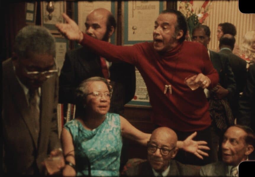 Still of the actors in Once Upon a Time in Harlem a man in a red sweater gestures toward a group of men and women gathered inside.